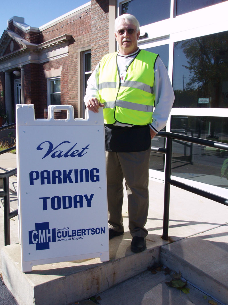 An older gentlemen standing beside a valet parking sign helping park vehicles