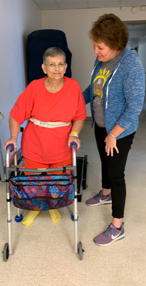 Photo of an older woman using a walker to walk while a care team employee assists her