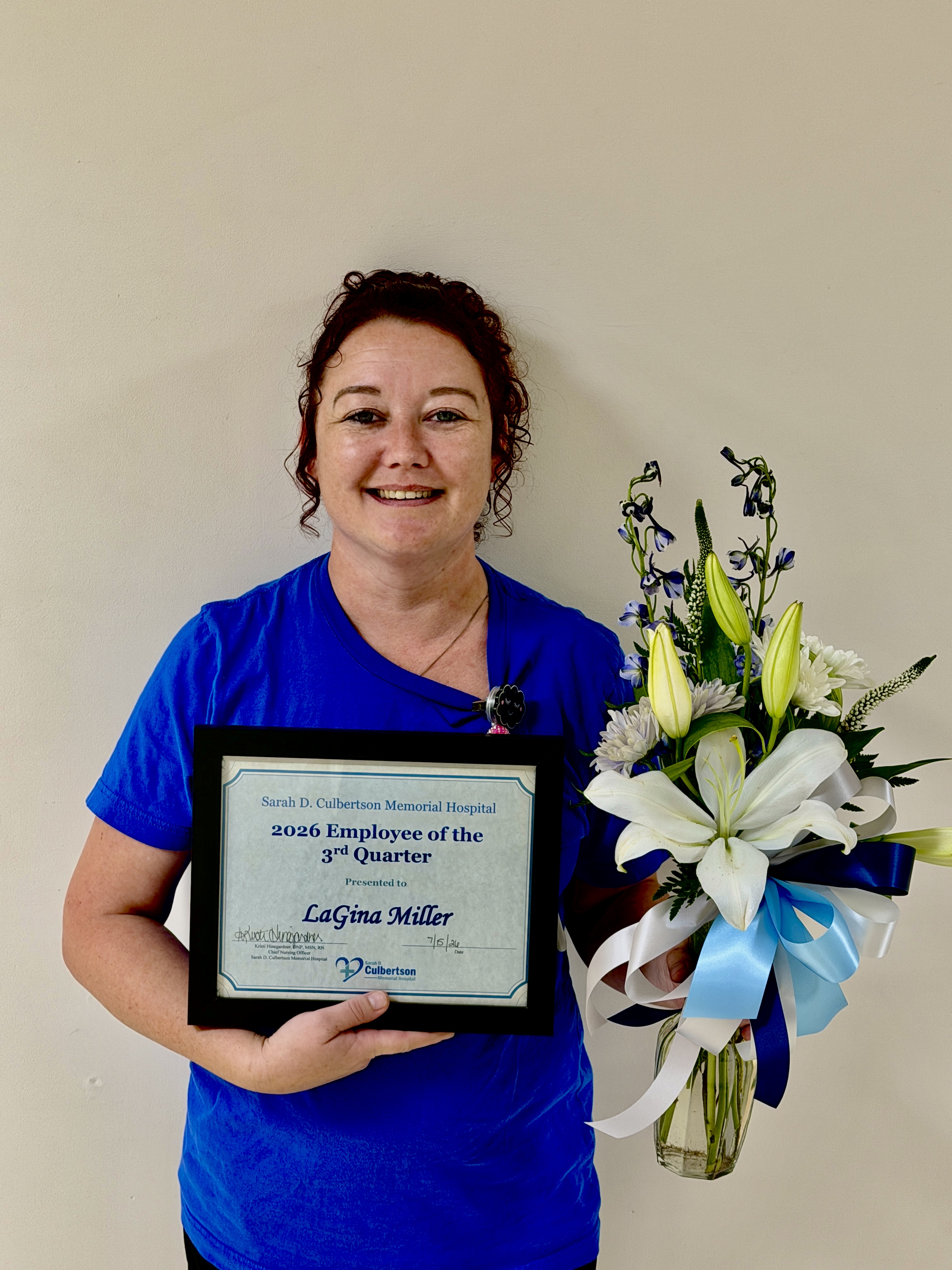 Curly haired woman with her hair up in blue scrub top holding flowers and a certificate.